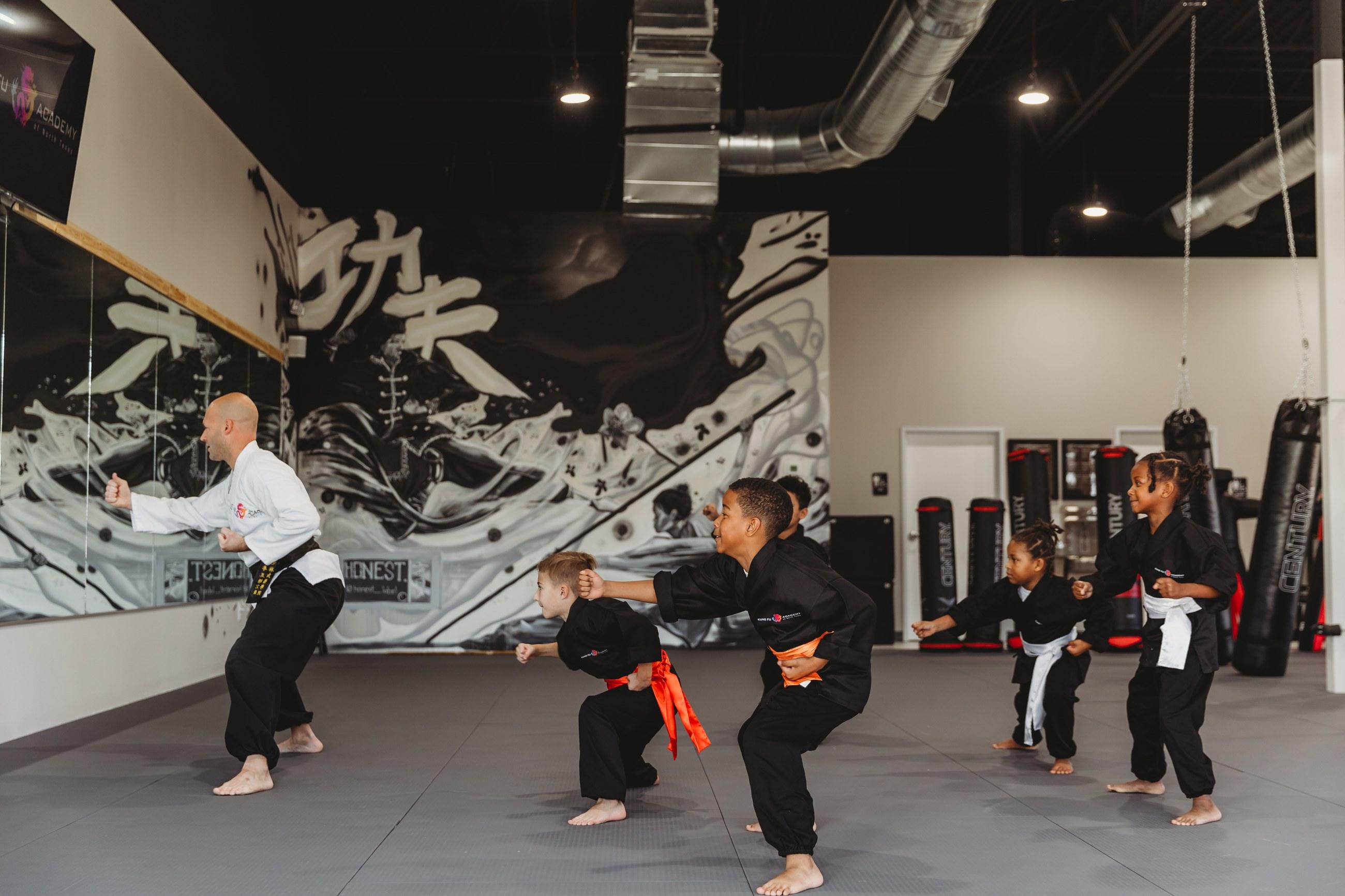 Instructor leading young students through a horse-stance drill in front of a hand-painted mural at Kung Fu Academy of North Texas, with heavy bags and an open ceiling, remodeled by NorthStar Commercial Interiors